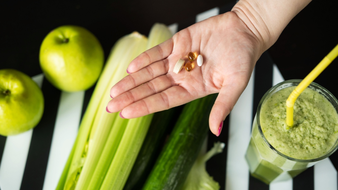 Woman holding detox supplements in the palm of her hand over various fruits and vegetables on a table